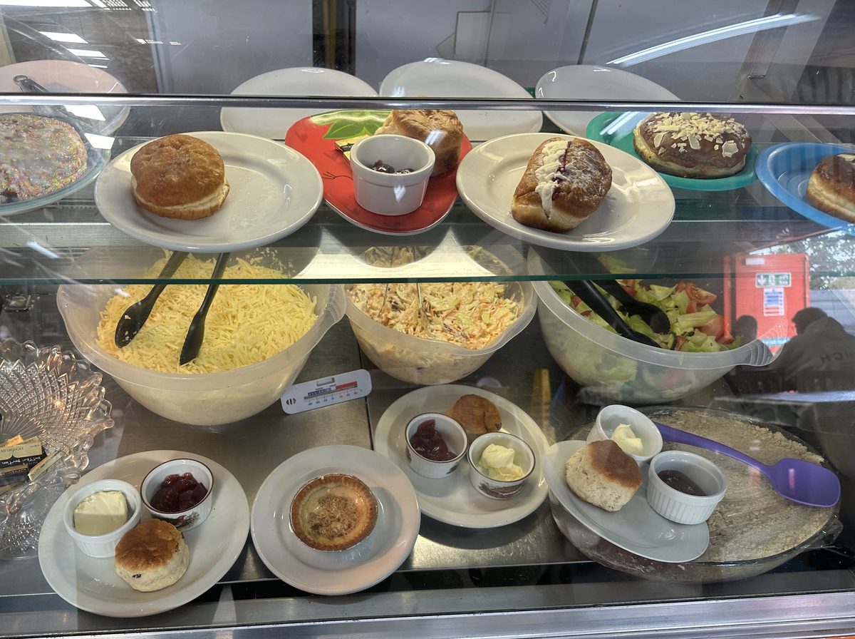 The Corner Café chilled cabinet, with scones set out with jam and clotted cream, doughnuts, a custard tart, and bowls of grated cheese, coleslaw and salad
