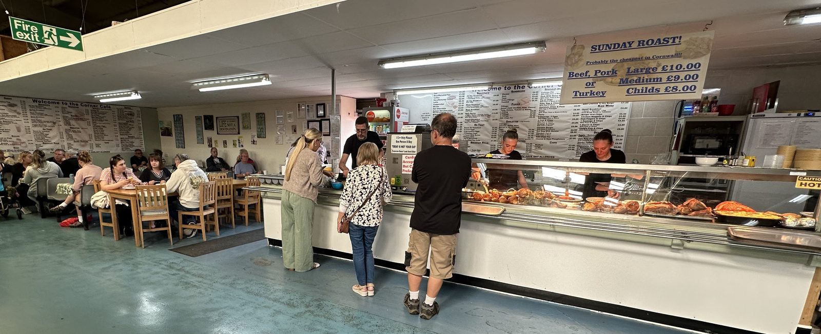 The full width of the Corner Café, with tables on both sides of the servery and the roast counter running along it, under the Sunday roast price banner