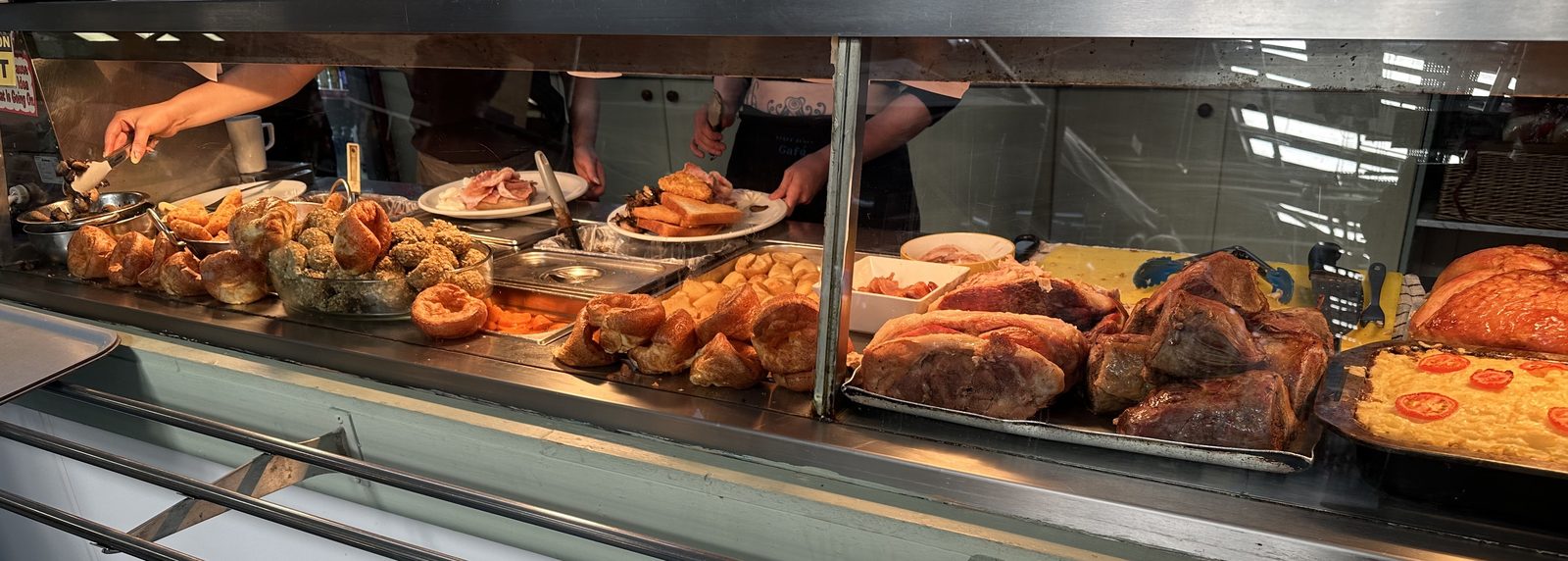 The Corner Café hot counter mid-service: trays of carved roast meat, Yorkshire puddings, roast potatoes, stuffing balls and a cheese and tomato flan, with staff plating up behind the glass