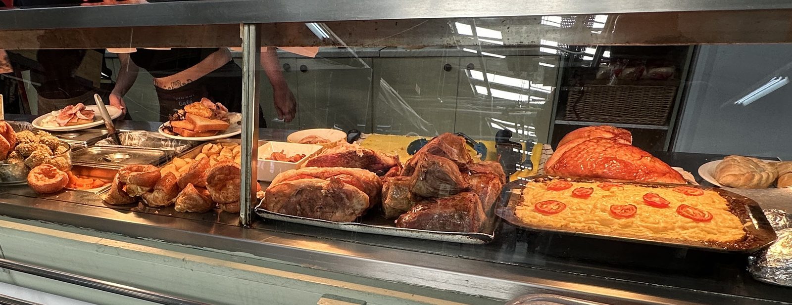 The Corner Café roast counter laid out for service: trays of carved roast meat, Yorkshire puddings, roast potatoes, stuffing balls and a cheese and tomato flan, with staff plating up behind the glass