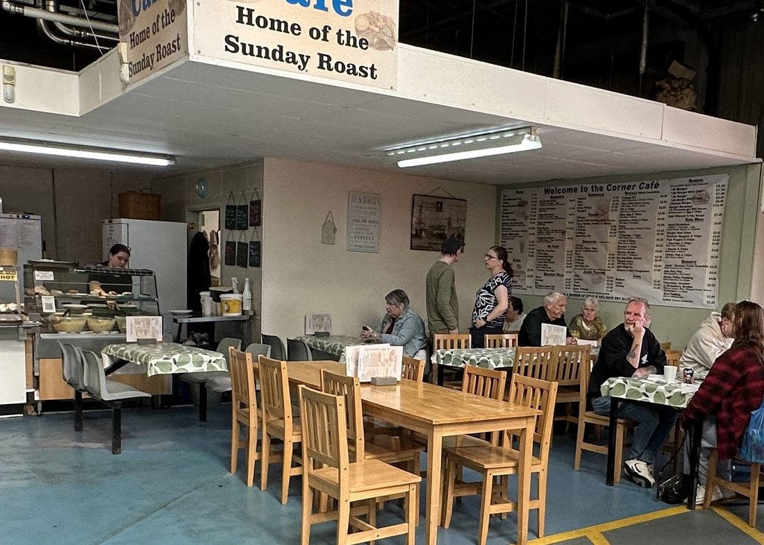 Families and market regulars sitting at the long wooden tables in the Corner Café, with the menu board on the wall behind them